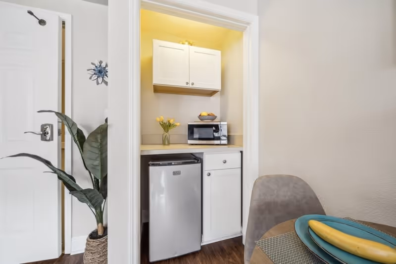 A small kitchenette area with a mini refrigerator, white cabinets, a microwave, and a vase with yellow flowers. To the right, part of a dining table with a banana on a blue plate and a gray chair are visible. A potted plant is placed near an open white door on the left.