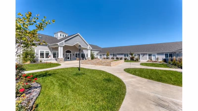 Front entrance of a single-story senior living building with a covered porte-cochere, curved concrete walkways, landscaped lawn, and clear blue sky.