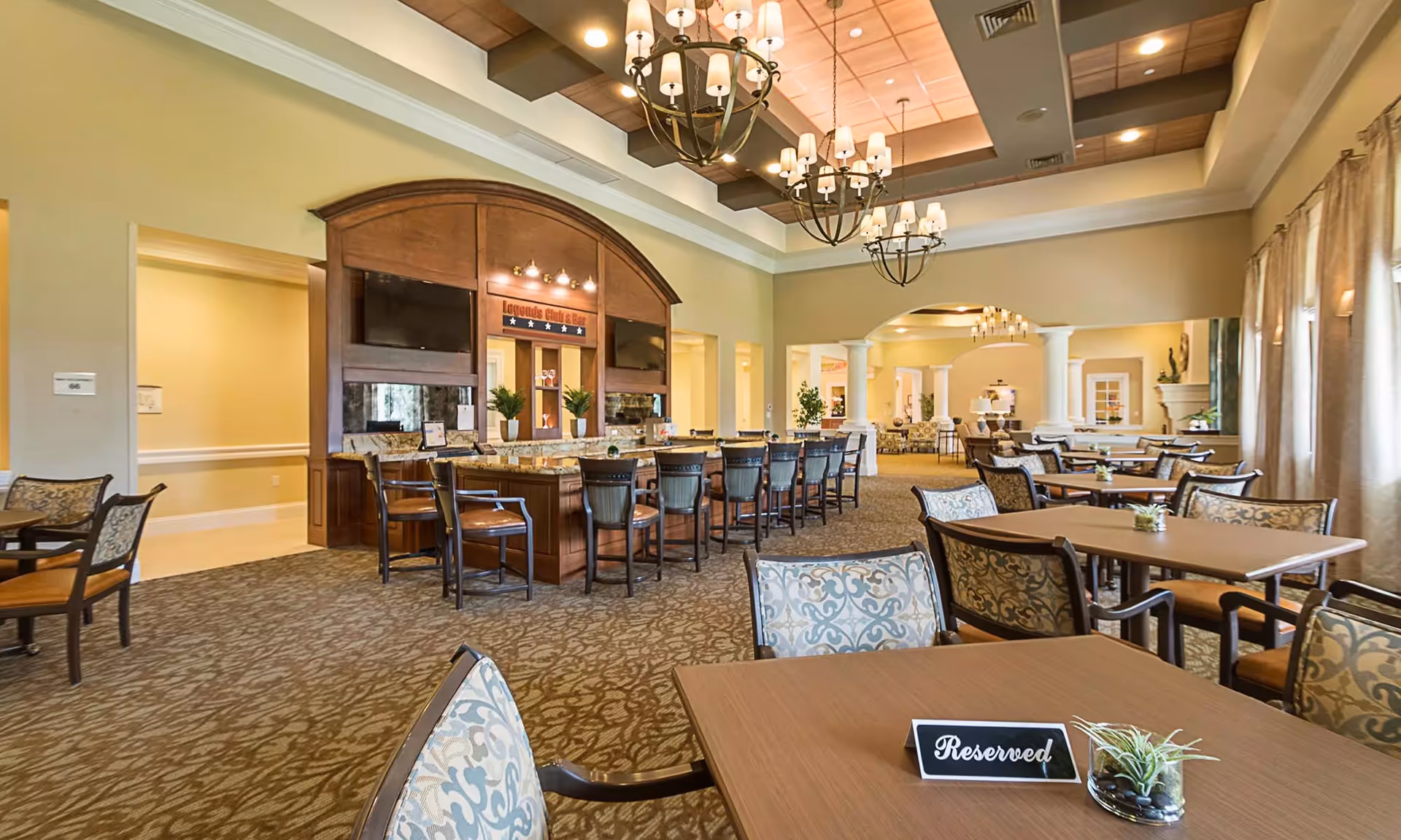 A spacious and elegant dining area in a senior living facility featuring multiple tables and chairs with patterned upholstery. A reserved sign is placed on the nearest table. The room has a high ceiling with decorative chandeliers and a wooden bar area labeled 'Legends Club & Bar' with bar stools. Large windows with curtains allow natural light to fill the room.