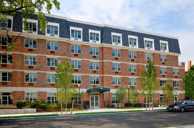 Exterior view of a multi-story brick building with numerous windows, a green awning above the entrance, small trees and shrubs in front, and a parked car on the street.