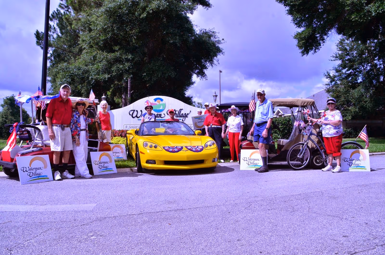 A group of senior adults standing outdoors in front of a sign that reads 'Waterman Village'. They are posing with a yellow convertible car and two golf carts decorated with American flags. The seniors are dressed casually and appear to be enjoying a sunny day.