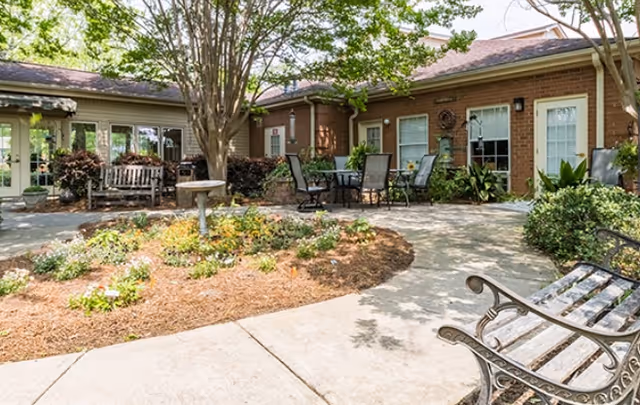 Outdoor courtyard area at Dublin Square featuring a circular garden bed with flowers and mulch, surrounded by a concrete walkway. There are several benches and a table with chairs under trees providing shade. The background shows a brick building with windows and doors.