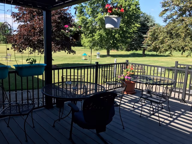 Outdoor patio area with metal tables and chairs under a covered structure, hanging flower pots, and a view of a grassy yard with trees and bird feeders in the background.