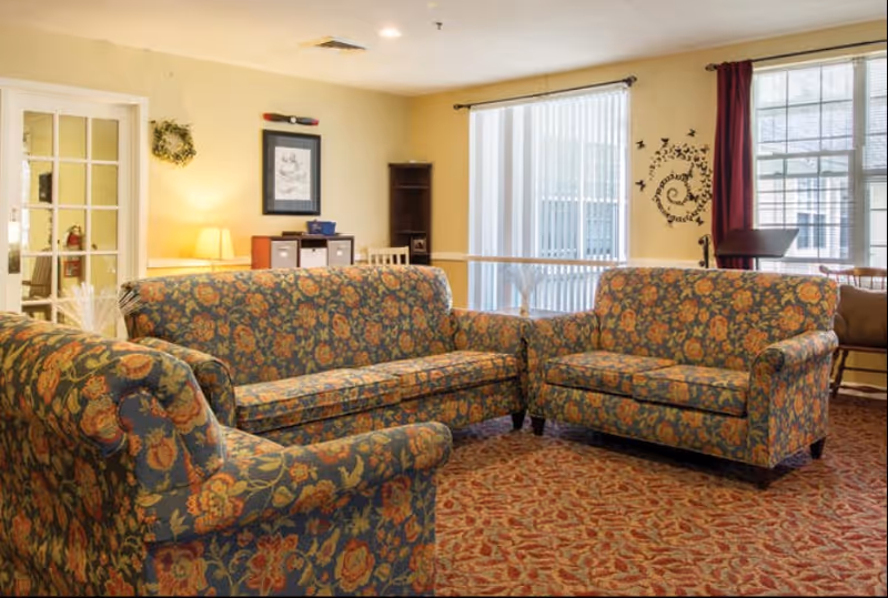 A cozy communal living room with floral-patterned sofas arranged on a patterned carpet near large windows.