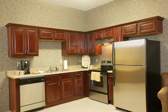 A kitchen area with dark wooden cabinets, a stainless steel refrigerator, stove, and dishwasher. The countertop has a coffee maker, paper towel holder, and some dishes. The walls have a patterned wallpaper and there is a beige armchair partially visible on the right.