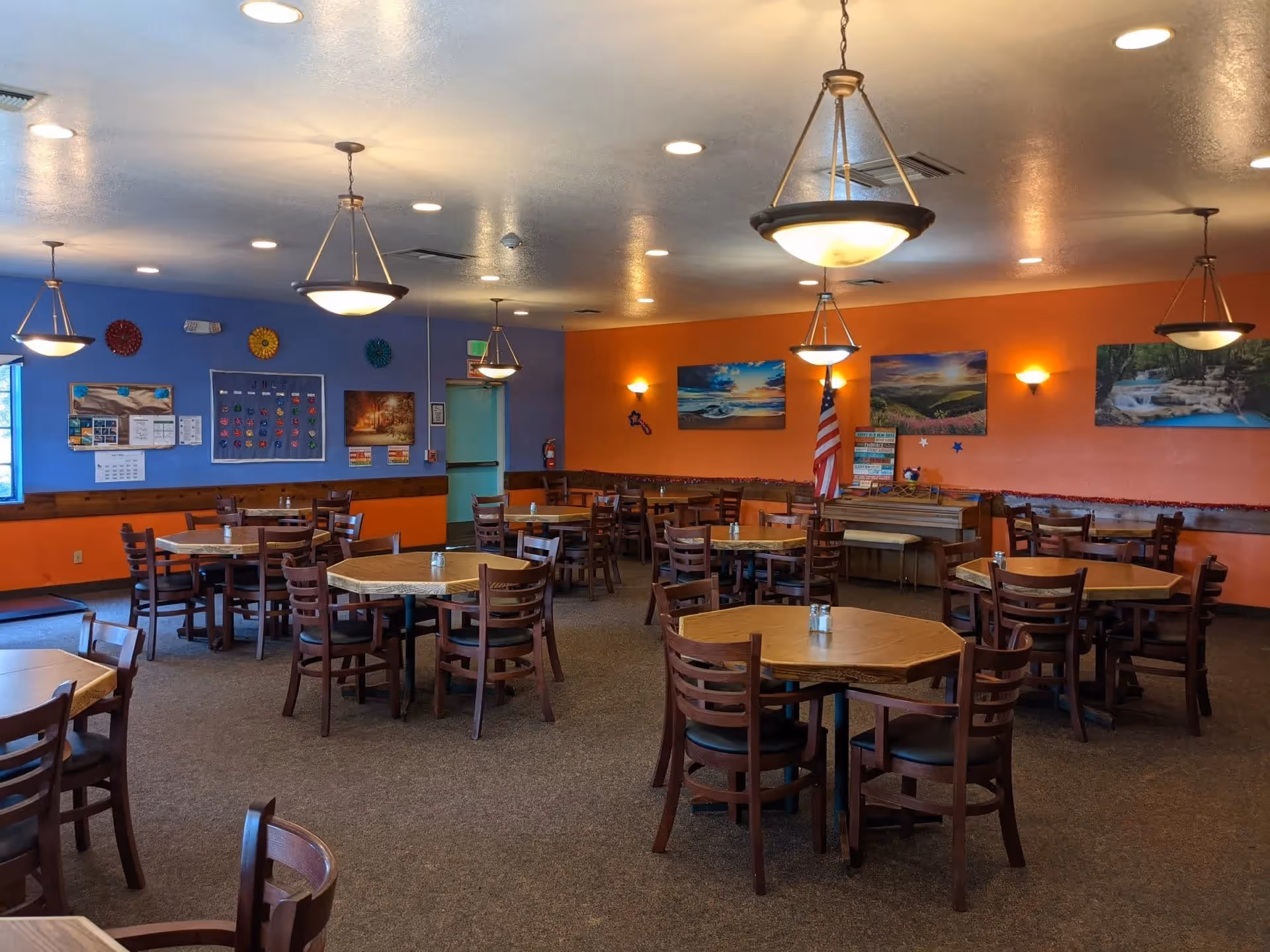 A spacious dining room with multiple wooden tables and chairs arranged neatly. The walls are painted orange and blue, decorated with landscape paintings and colorful wall hangings. Several ceiling lights hang from the ceiling, illuminating the room. An American flag is displayed near the back wall.
