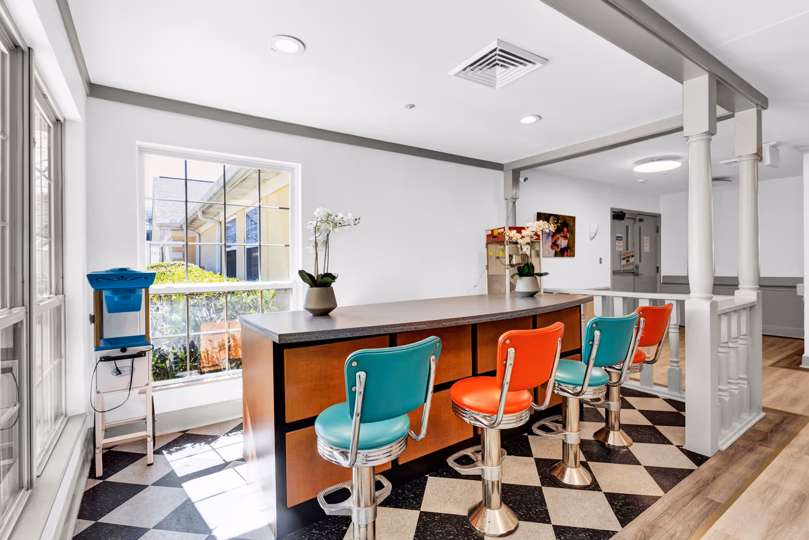 Bright interior space with a counter and four retro-style bar stools in alternating teal and orange colors. The floor has a black and white checkered pattern near the counter and wood flooring beyond. Large windows allow natural light to fill the room, and there are two potted orchid plants on the counter. The area appears clean and welcoming with white walls and ceiling lights.