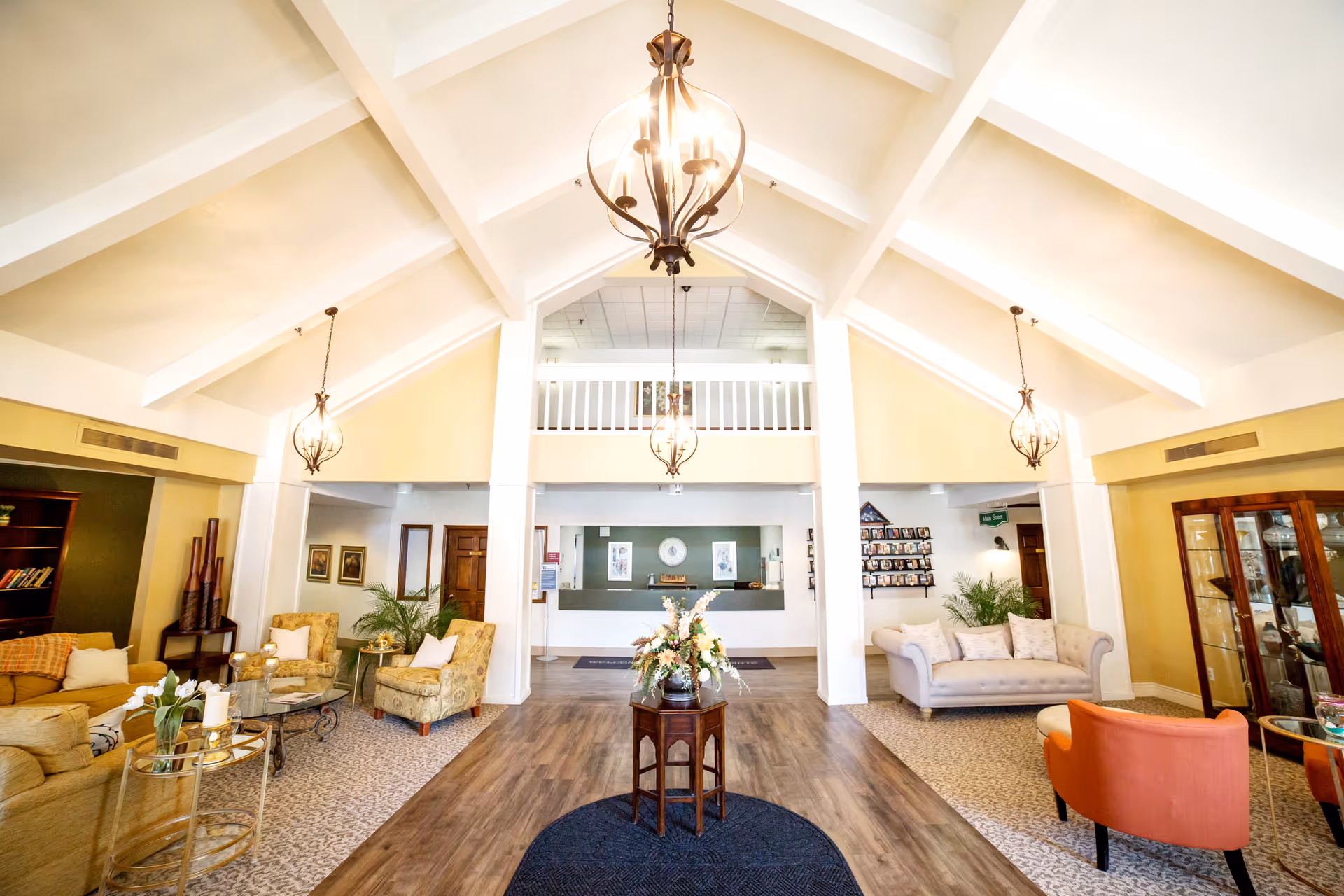 Bright and spacious retirement community lobby with high vaulted ceilings and three hanging chandeliers. The room features comfortable seating areas with sofas and armchairs, a small table with a floral arrangement in the center, and a reception desk at the back. The decor includes plants, framed pictures, and a display cabinet.