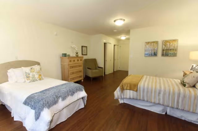A spacious bedroom with two beds, one on the left with white bedding and a blue throw blanket, and one on the right with striped bedding and a mustard-colored throw blanket. There is a wooden dresser with decorative items and a green armchair against the wall. The room has wooden flooring, two framed pictures on the wall, and ceiling lights.