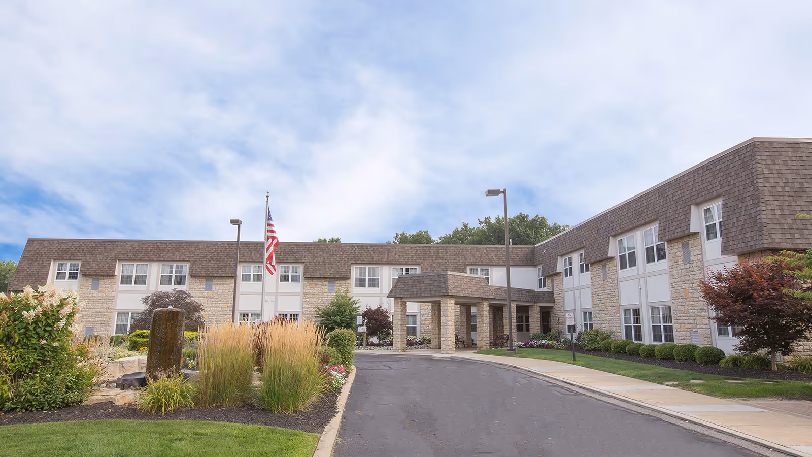 Exterior view of a two-story senior living facility building with stone and siding facade, a covered entrance, landscaped garden with ornamental grasses and shrubs, an American flag on a flagpole, and a paved driveway under a partly cloudy sky.