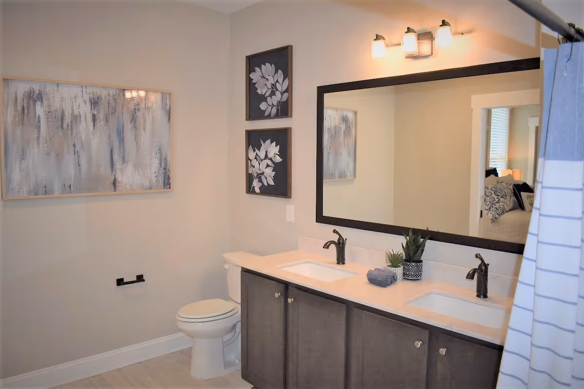 A modern bathroom featuring a double sink vanity with dark cabinets and two black faucets. Above the vanity is a large rectangular mirror with a black frame and a three-light fixture. To the left of the vanity is a white toilet, and above it are two framed botanical prints. A large abstract painting hangs on the adjacent wall. The bathroom has light-colored walls and flooring, and a shower curtain is partially visible on the right side.