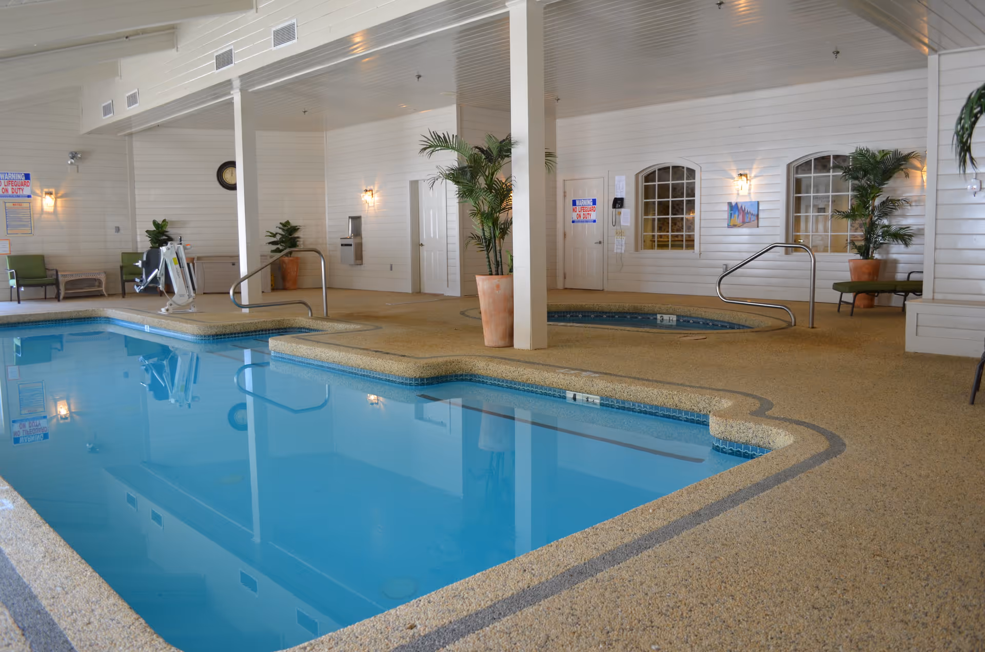 Indoor swimming pool area with clear blue water, surrounded by a textured beige floor. There are handrails for pool access, potted plants, and seating areas with green cushioned chairs. The walls are white with windows and wall-mounted lights, and there is a hot tub in the background.
