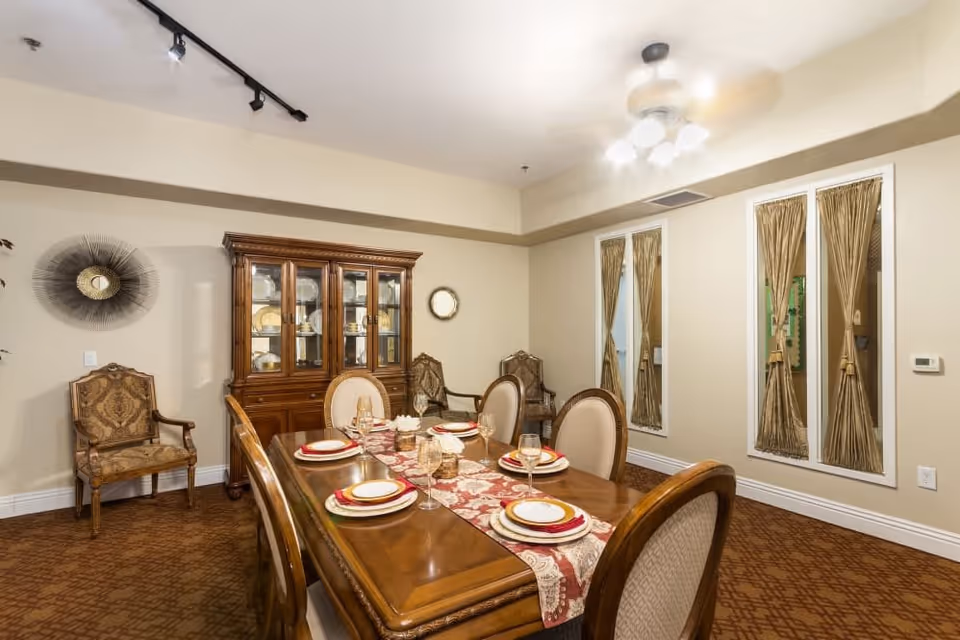 A formal dining room with a wooden dining table set for six people with plates, glasses, and a decorative table runner. The room features upholstered chairs, a wooden china cabinet displaying dishes, two decorative wall mirrors with gold curtains, and a ceiling fan with lights. The walls are painted beige and the floor is carpeted with a patterned design.