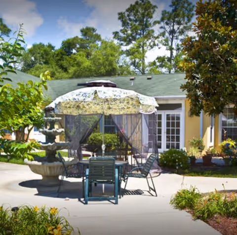 Courtyard patio with a table and chairs under a floral umbrella canopy beside a tiered fountain in front of a light-yellow building.