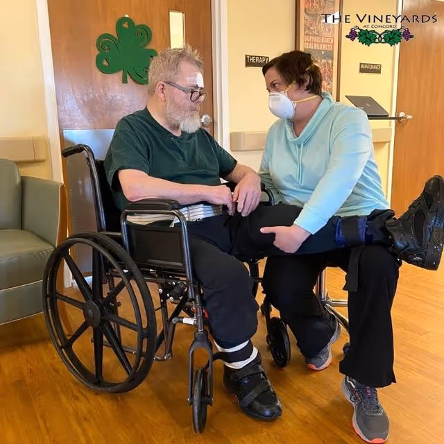 A man in a wheelchair with a bandage on his forehead is receiving physical therapy from a woman wearing a face mask. The woman is holding and lifting the man's leg while they look at each other in a room with wooden floors and doors labeled 'Therapy' and 'Maintenance'.