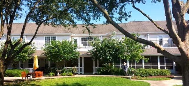 Two-story senior living facility building with a covered porch, surrounded by green trees and bushes. There is a wooden bench and table with a yellow umbrella on the left side of the image, and a person standing near the entrance. The sky is clear and blue.