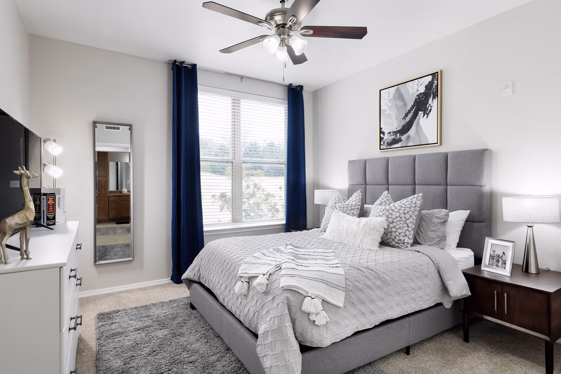 A modern bedroom with a gray upholstered bed featuring multiple decorative pillows and a textured blanket. There is a dark wooden nightstand with a lamp and a framed photo on the right side of the bed. A large window with white blinds and dark blue curtains is behind the bed. On the left side, there is a white dresser with a gold giraffe figurine, books, and a TV. A full-length mirror is mounted on the wall next to the dresser. The room has a ceiling fan with lights and light-colored walls and carpet.