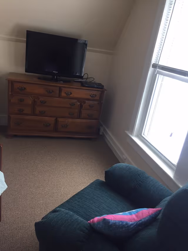 A cozy room corner featuring a wooden dresser with multiple drawers, a flat-screen TV on top, a remote control device, a window with blinds partially open, and a dark green armchair with a colorful pillow.