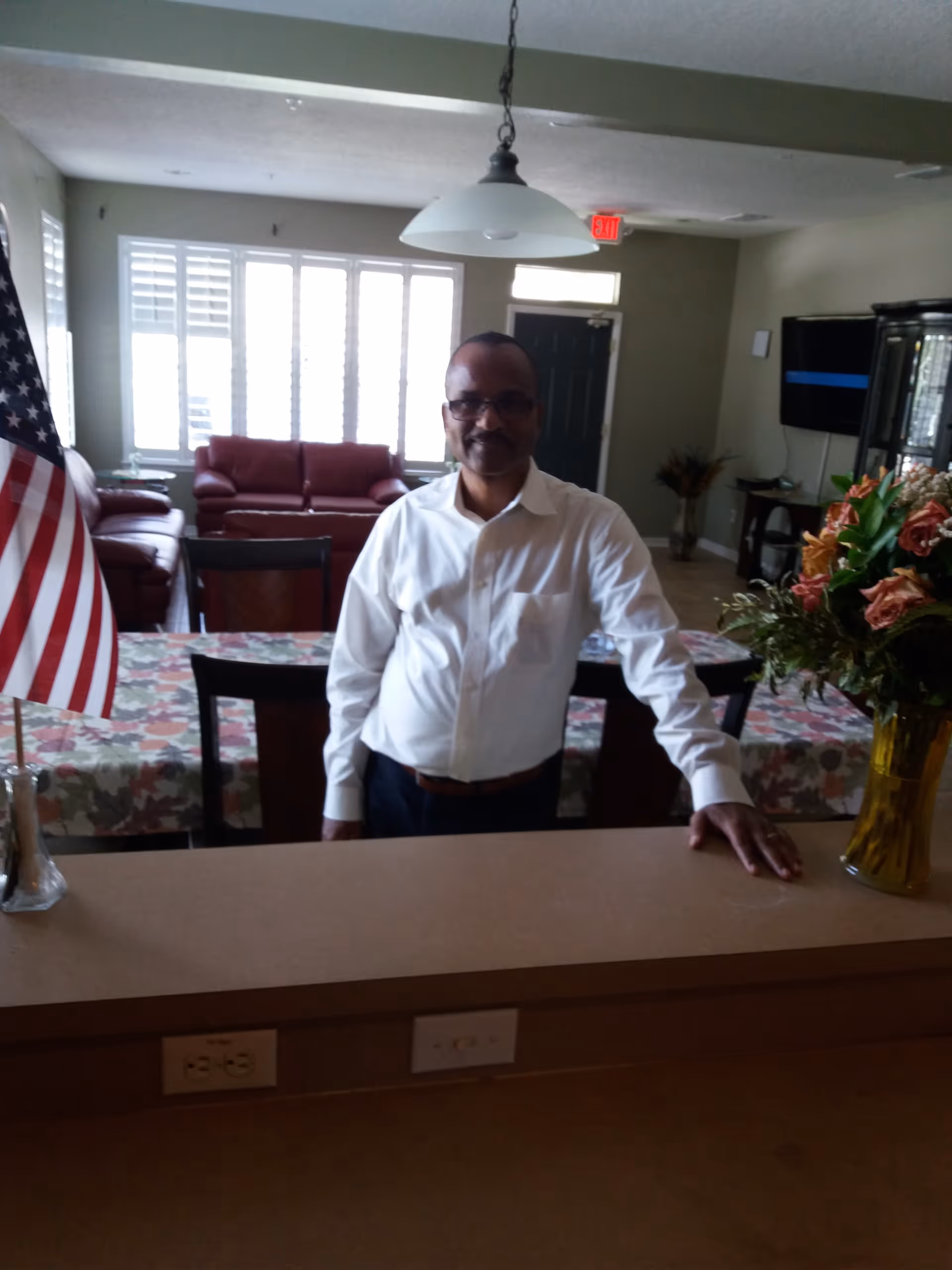 A man wearing glasses and a white shirt stands behind a counter in a living room area. The room has a table with a floral tablecloth, chairs, a vase with flowers, an American flag, a red sofa, a TV mounted on the wall, and a door with an exit sign above it.