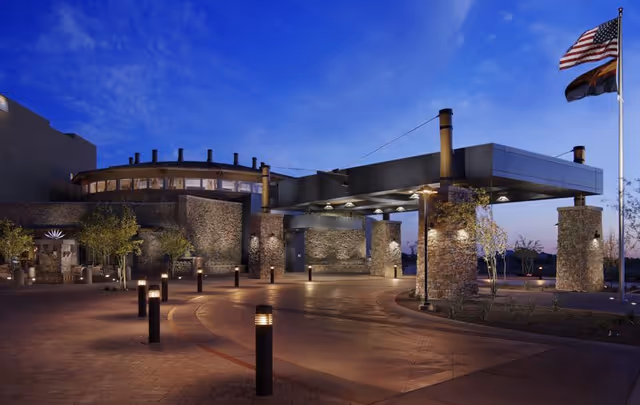 Stone and metal entrance of a senior living facility at dusk with a covered porte-cochere, illuminated bollards, and flagpoles.