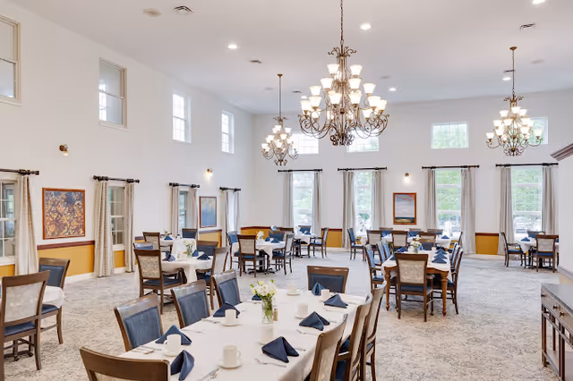 A spacious dining room with multiple tables set with white tablecloths, navy blue folded napkins, white cups, and small flower arrangements. The room has large windows with curtains, several chandeliers hanging from the ceiling, and framed artwork on the walls. The carpet is light-colored with a subtle pattern.