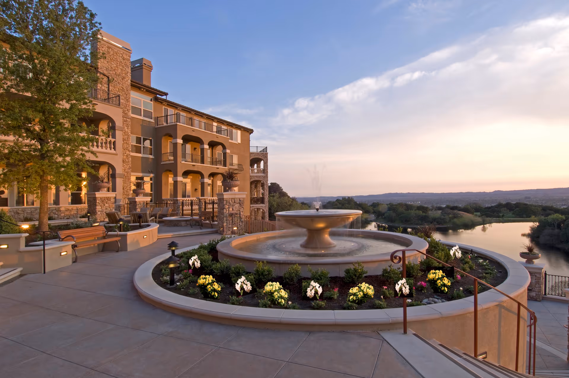 Outdoor view of a senior living facility at sunset featuring a large circular fountain surrounded by flower beds and a paved walkway. The building has multiple balconies and stone accents, with benches and trees nearby. A river and distant hills are visible in the background under a partly cloudy sky.