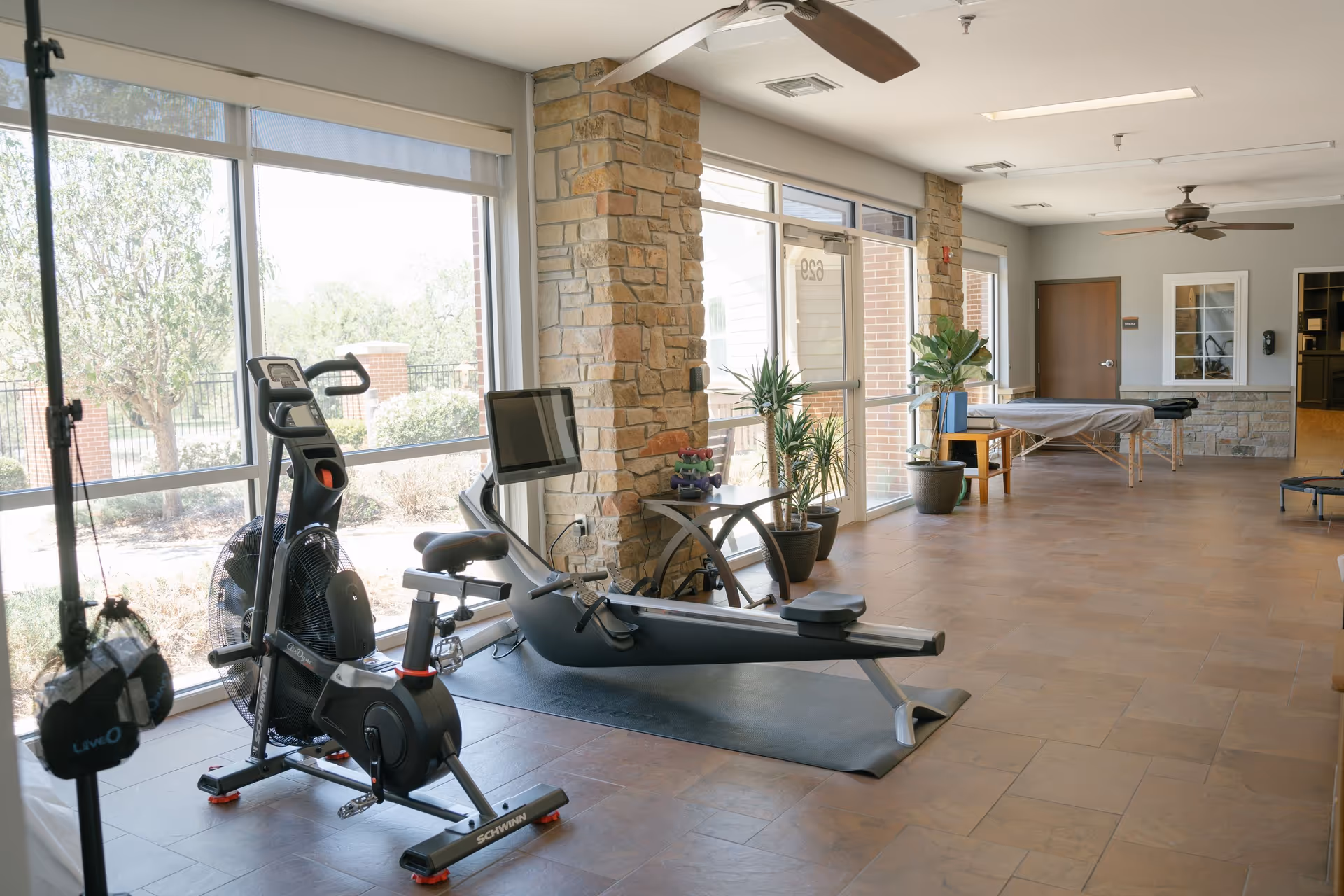 A bright exercise room with large windows showing outdoor greenery. Inside, there is a stationary exercise bike, a rowing machine, several potted plants, and a massage table. The room has tiled floors, stone pillars, ceiling fans, and a door leading outside.