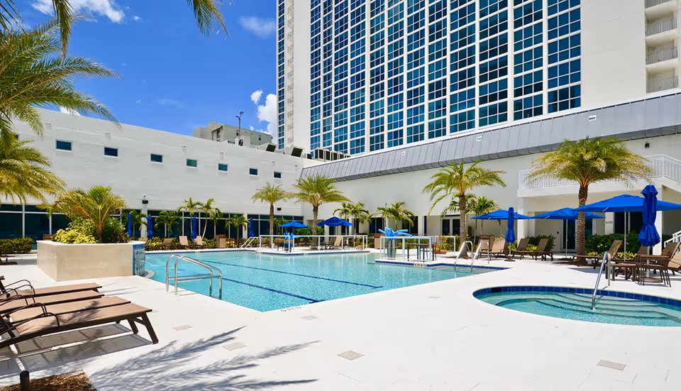 Outdoor swimming pool area at The Palms of Fort Myers with lounge chairs, palm trees, blue umbrellas, and a hot tub under a clear blue sky. A tall building with many windows is visible in the background.
