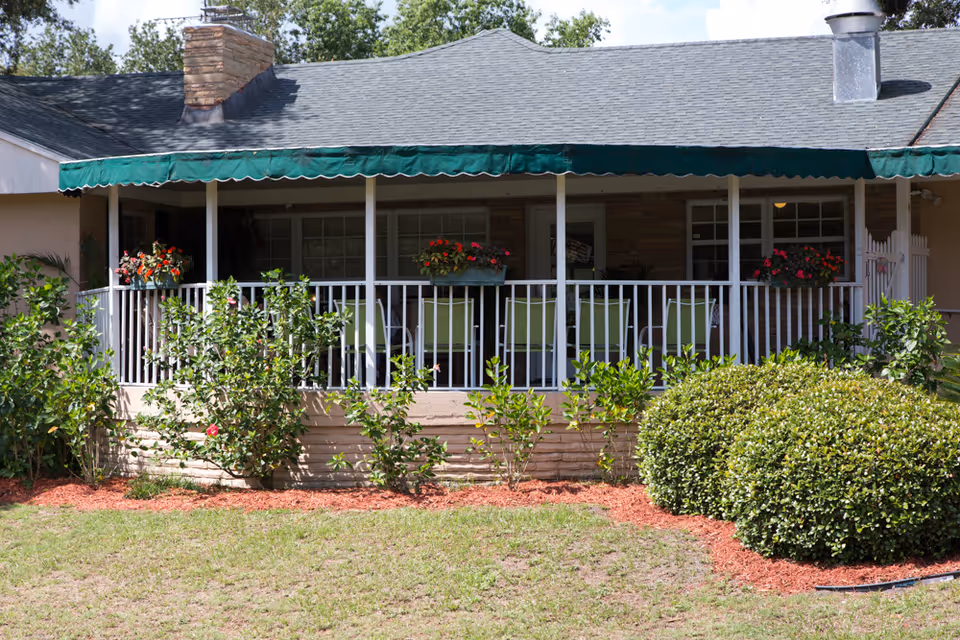 Front view of a single-story building with a covered porch featuring green chairs and flower pots. The porch has a white railing and a green awning. There are bushes and plants in front of the porch with a grassy lawn in the foreground.
