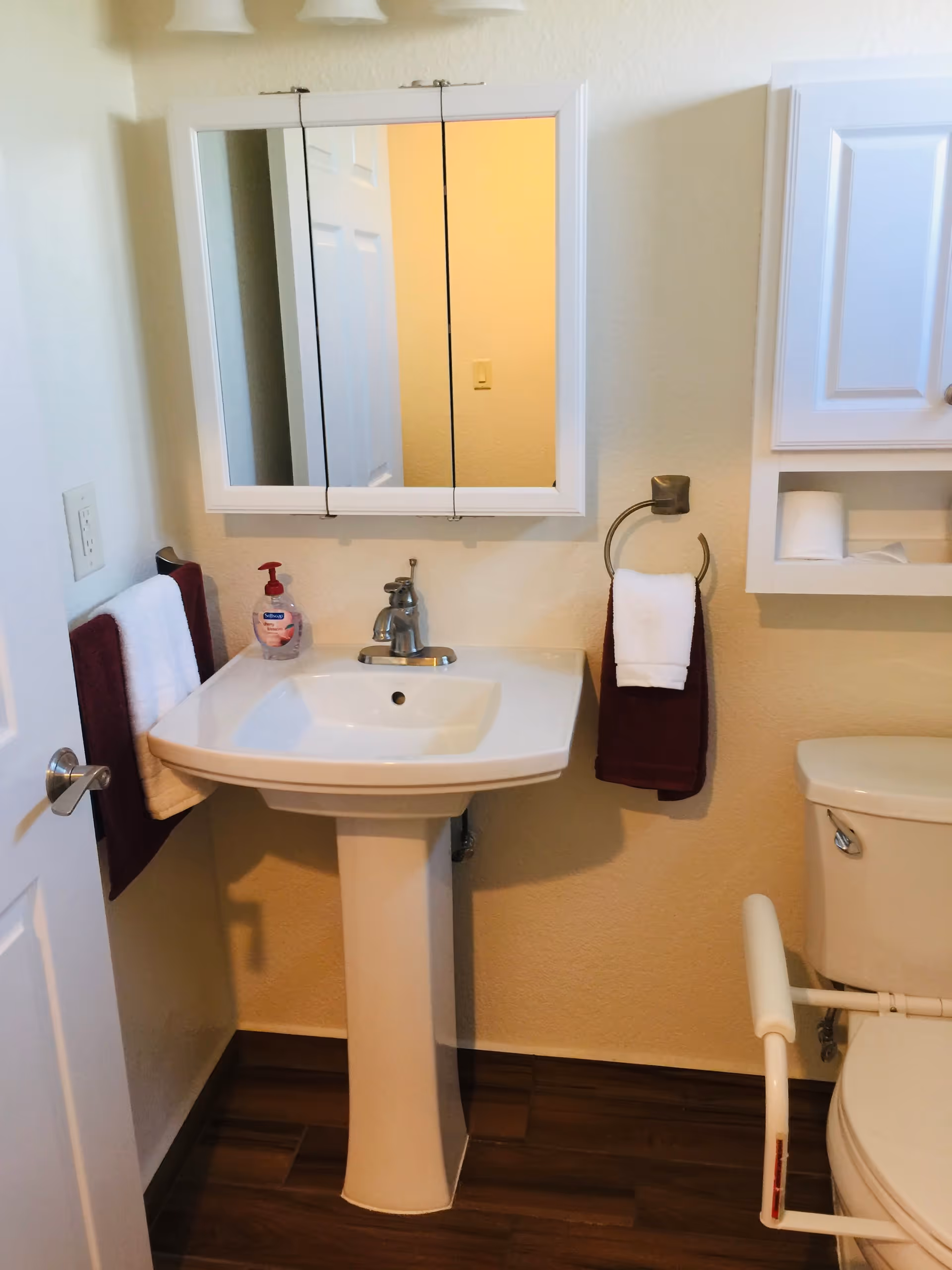 A small bathroom with a white pedestal sink, a wall-mounted mirror cabinet above the sink, a toilet with a safety rail, and two towel holders with burgundy and white towels. There is a soap dispenser on the sink and a wall cabinet with toilet paper above the toilet.