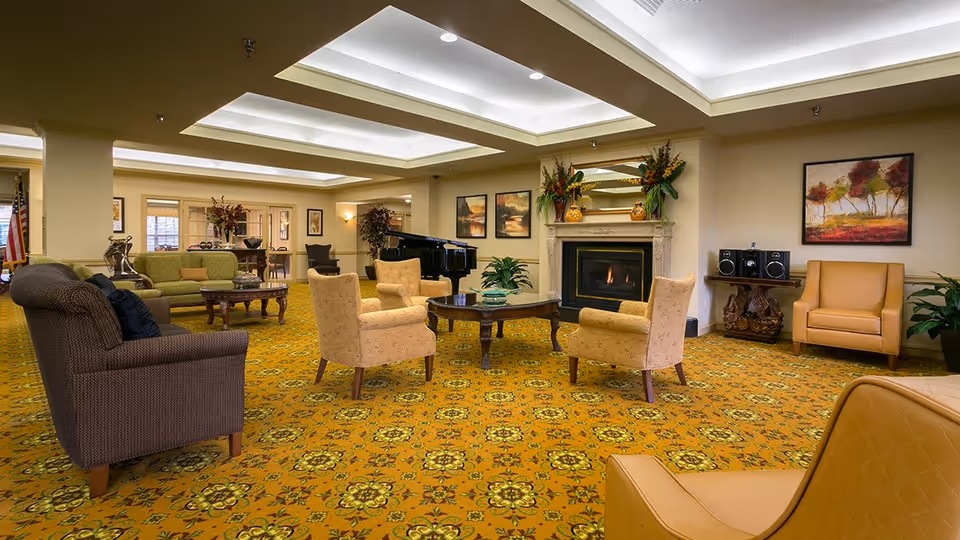 A spacious senior living community lounge with patterned yellow carpet, multiple armchairs and sofas arranged around a central coffee table, a fireplace with decorative plants and a mirror above it, framed artwork on the walls, and a grand piano in the background.