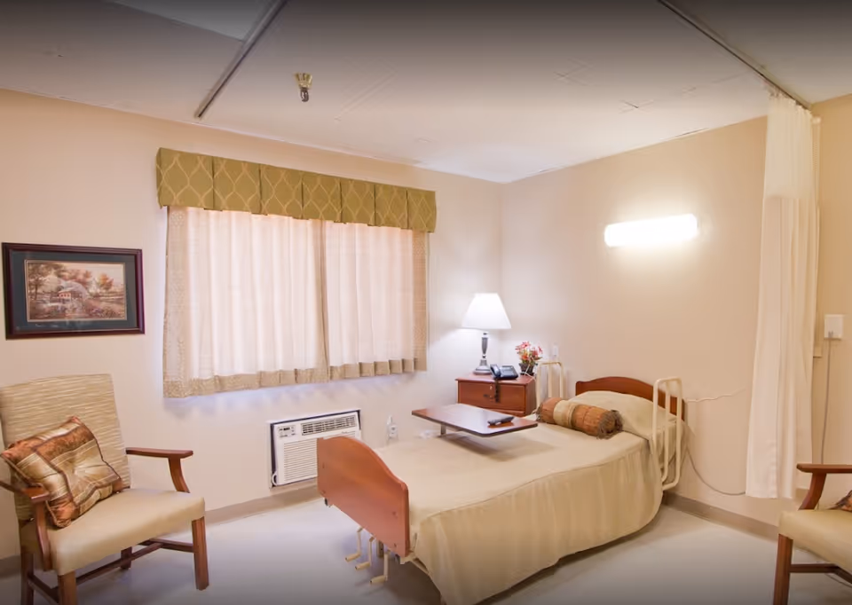 A simple senior living facility bedroom with a single bed covered in beige bedding, a wooden bedside table with a lamp, a telephone, and a small flower arrangement. There is a beige armchair with a decorative pillow, a window with curtains and a green valance, and a wall-mounted air conditioning unit below the window. A privacy curtain is partially visible on the right side of the room.