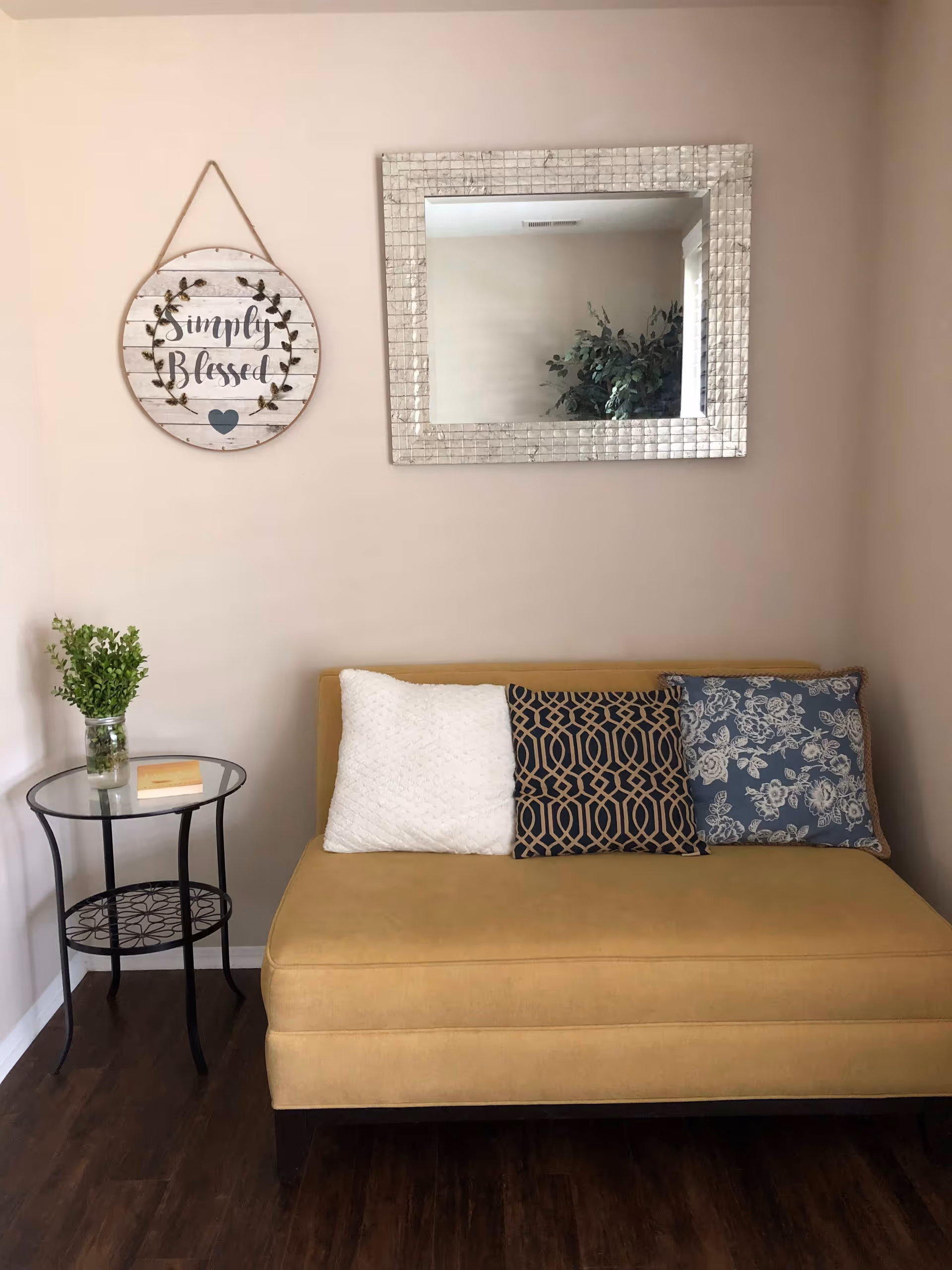 A cozy seating area with a mustard yellow cushioned bench adorned with three decorative pillows in white, navy blue with a geometric pattern, and blue with floral designs. Above the bench is a large rectangular mirror with a textured frame reflecting a plant and a window. To the left of the bench is a small round glass-top side table with a black metal frame, holding a small green plant in a glass jar and a book. On the wall above the table hangs a round wooden sign with the words 'Simply Blessed' surrounded by a leafy wreath design.