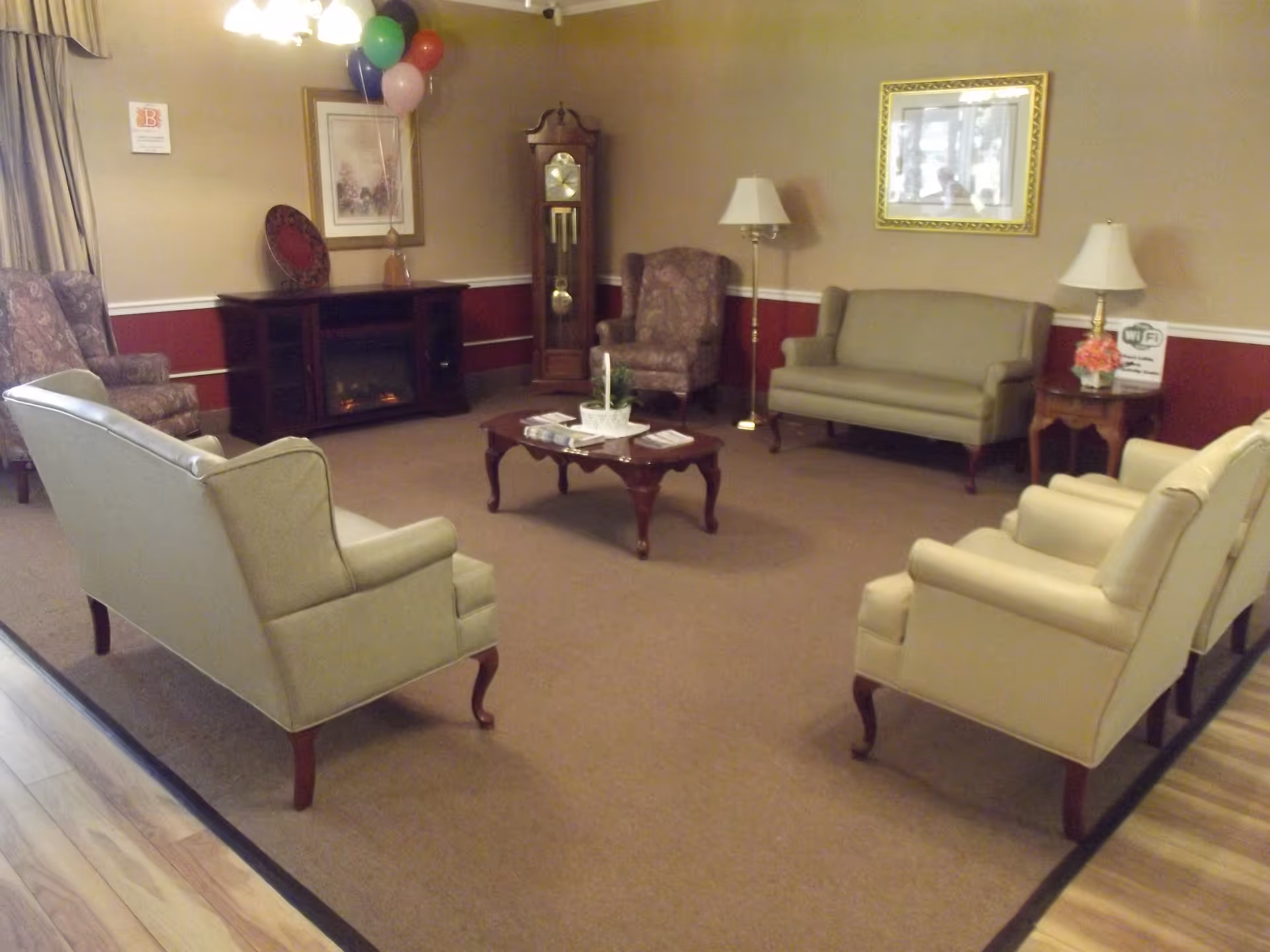 A cozy living room area with beige and patterned armchairs, two beige sofas, a wooden coffee table with magazines and a plant, a grandfather clock, a fireplace, two floor lamps, a side table with a lamp and flowers, and framed artwork on the walls. Balloons are tied near the fireplace.