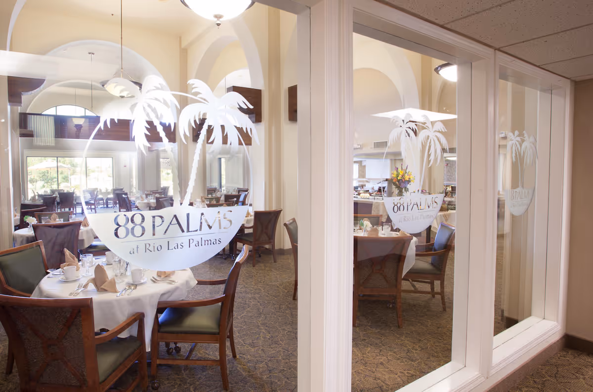 View through glass doors into a dining room with tables set with white tablecloths, napkins, cups, and silverware. The glass doors have a white decal logo with two palm trees and the text '88 PALMS at Rio Las Palmas'. The dining room has wooden chairs with green cushions and large windows letting in natural light.