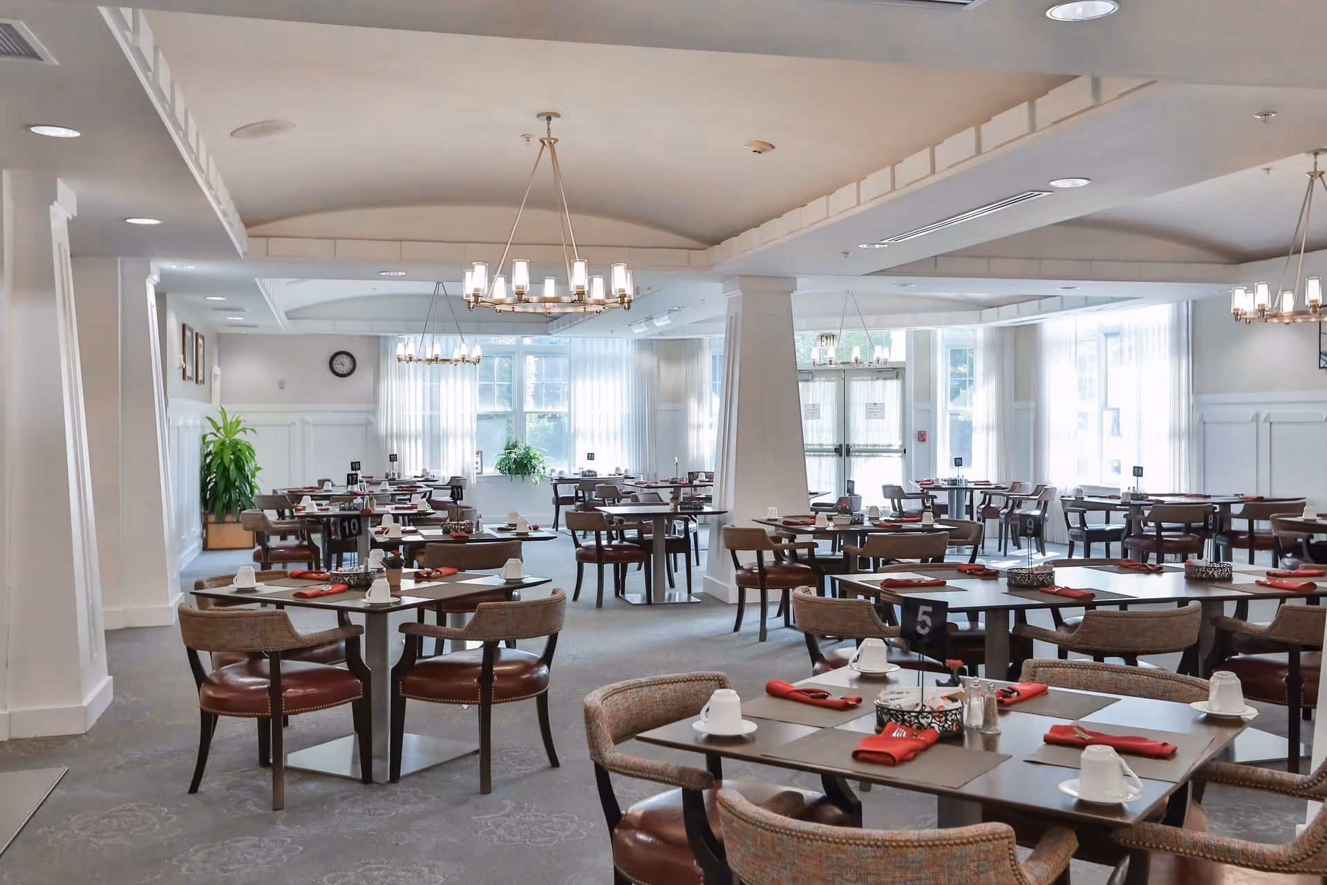 A bright and spacious dining room with multiple tables and chairs arranged neatly. Each table is set with white cups, red napkins, and small condiment containers. Large windows with sheer white curtains allow natural light to fill the room. Modern chandeliers hang from the ceiling, and there are green plants placed near the walls.
