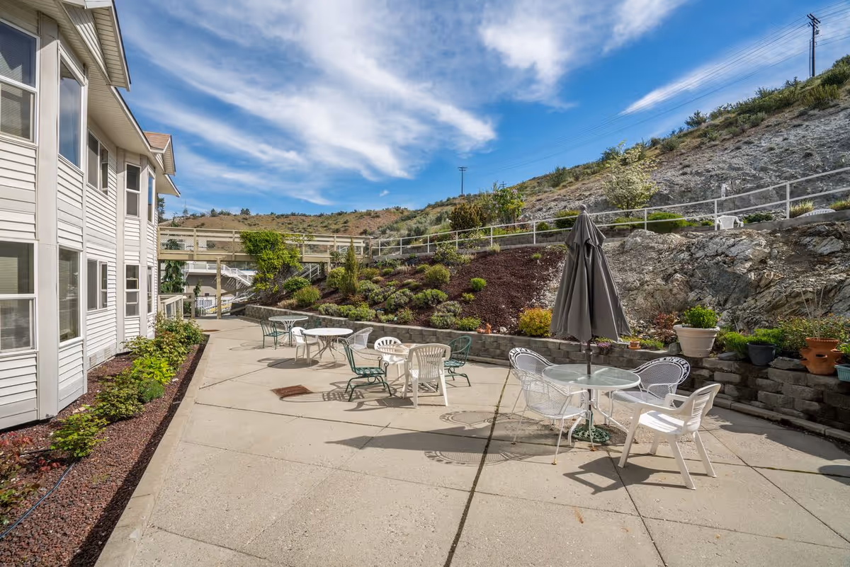 Outdoor patio area at Heritage Heights featuring several round glass and plastic tables with chairs arranged around them. One table has a closed umbrella. The patio is adjacent to a white building with multiple windows and is bordered by a landscaped hillside with plants and rocks under a partly cloudy blue sky.