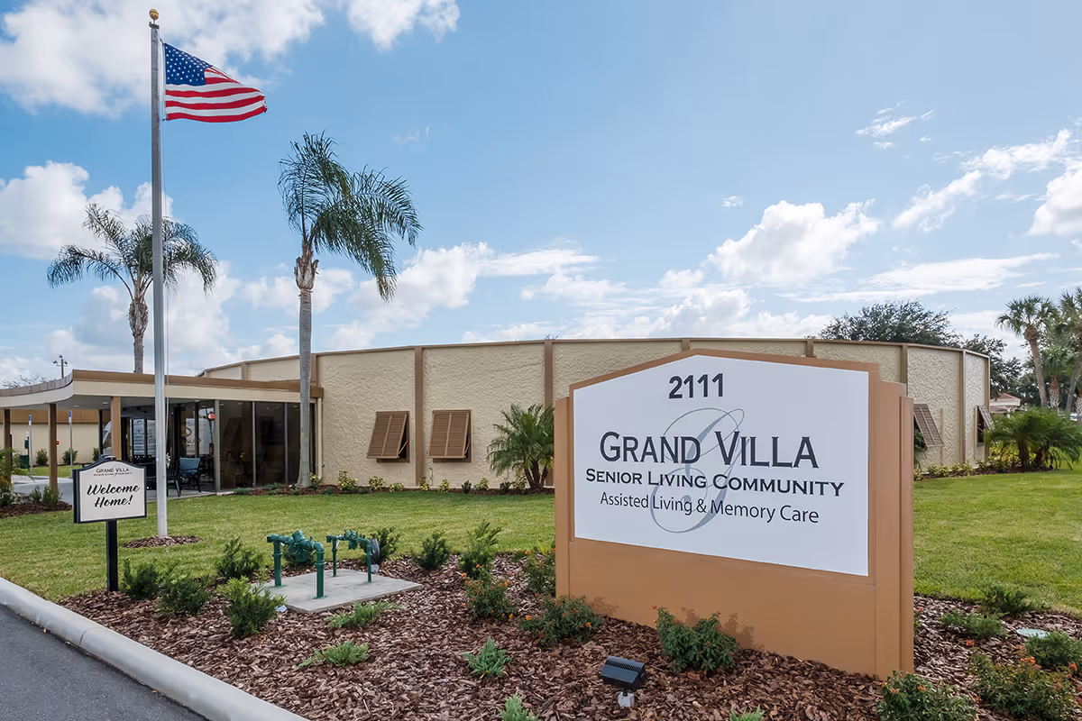 Exterior view of Grand Villa Senior Living Community building with a large sign displaying the name and services offered. The building is surrounded by well-maintained landscaping, palm trees, and an American flag on a flagpole. The sky is partly cloudy.