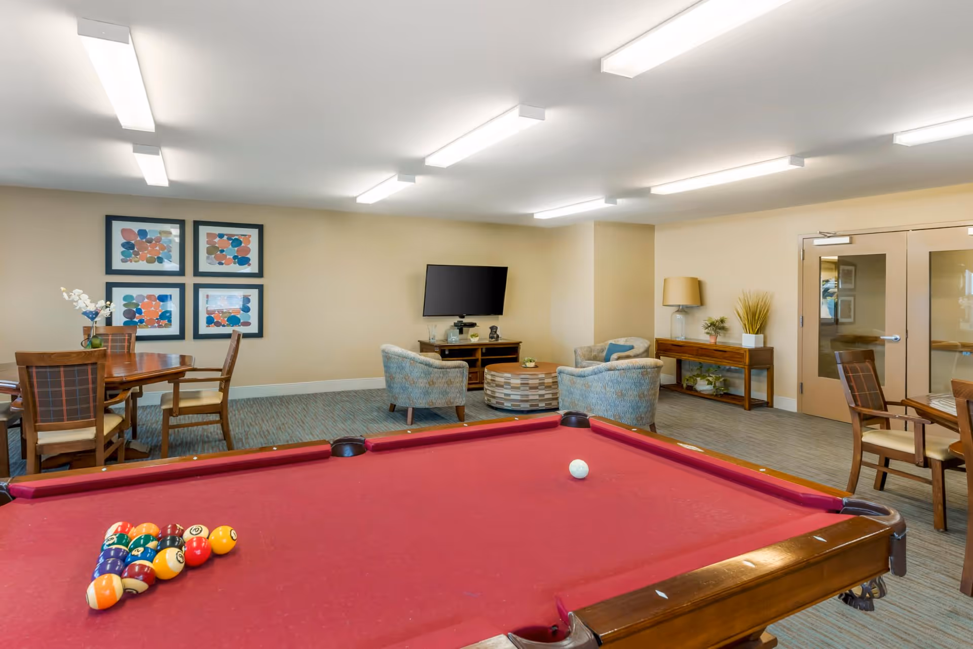 Community game room featuring a red pool table in the foreground, seating area with a TV, and a dining table in the background.