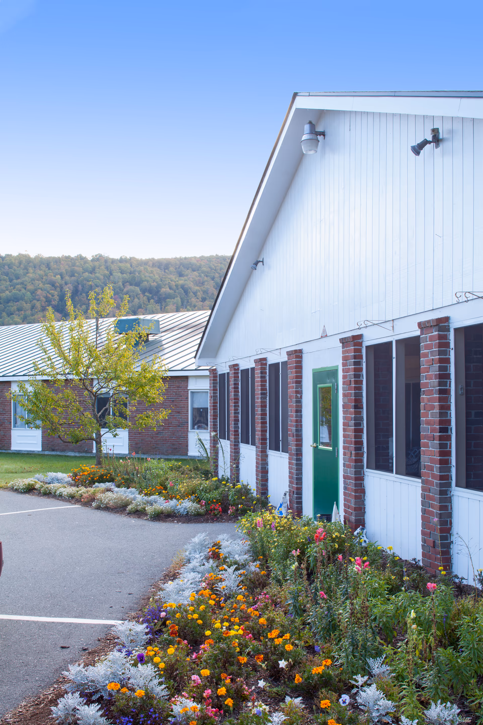 Exterior view of a building with white siding and brick columns, featuring a green door and multiple windows. A colorful flower bed with various blooming flowers lines the walkway in front of the building. In the background, there is another brick building and a tree with green leaves, with forested hills under a clear blue sky.