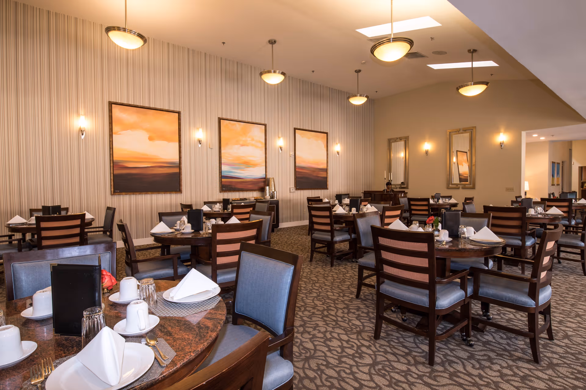 A spacious dining room with multiple round tables set with white plates, cups, napkins, and silverware. The room features patterned carpet, striped wallpaper, and three large paintings with sunset scenes on the wall. There are several pendant lights hanging from the ceiling and wall sconces providing warm lighting. Chairs with wooden frames and blue cushions surround the tables.