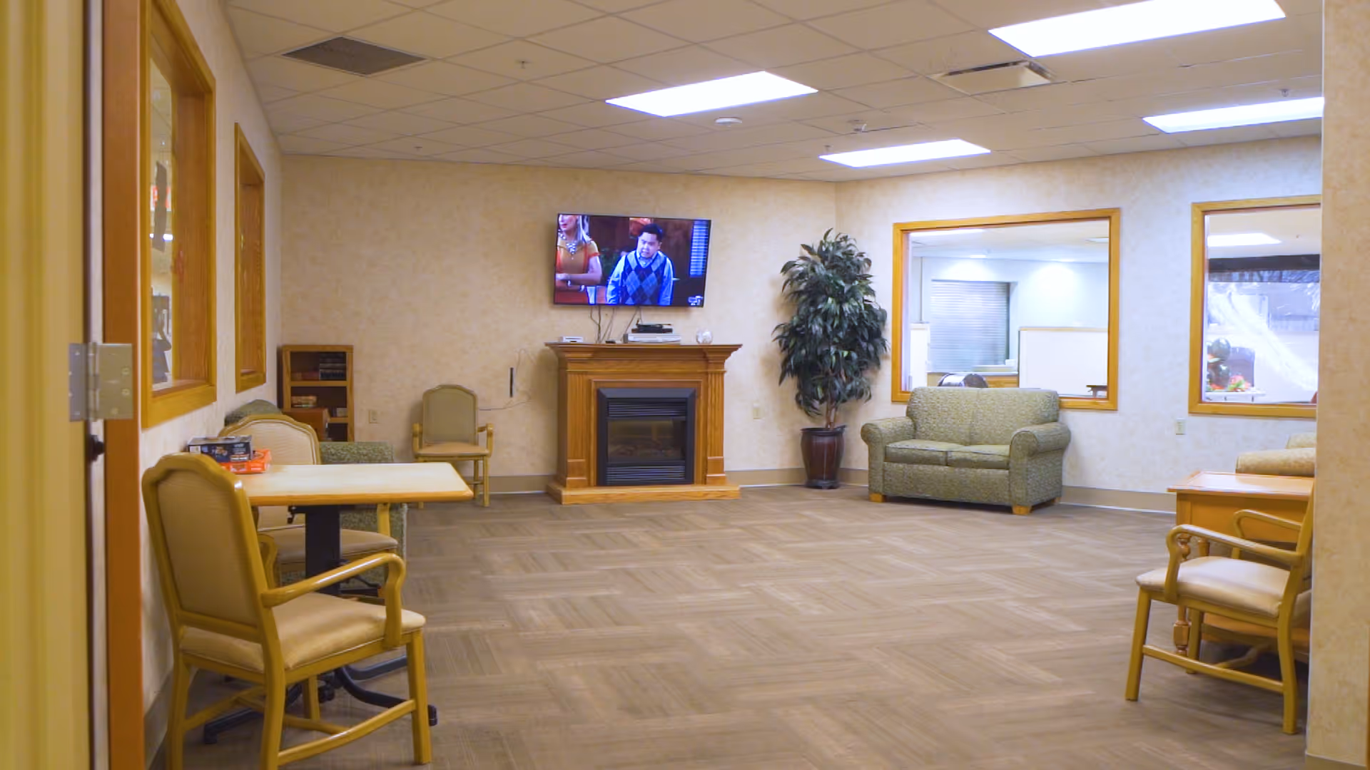 A spacious common area in a senior living facility with beige walls and carpeted floor. The room features a wall-mounted TV above a wooden electric fireplace, a green loveseat, several chairs, tables, and a large potted plant. Large windows look into adjacent rooms.