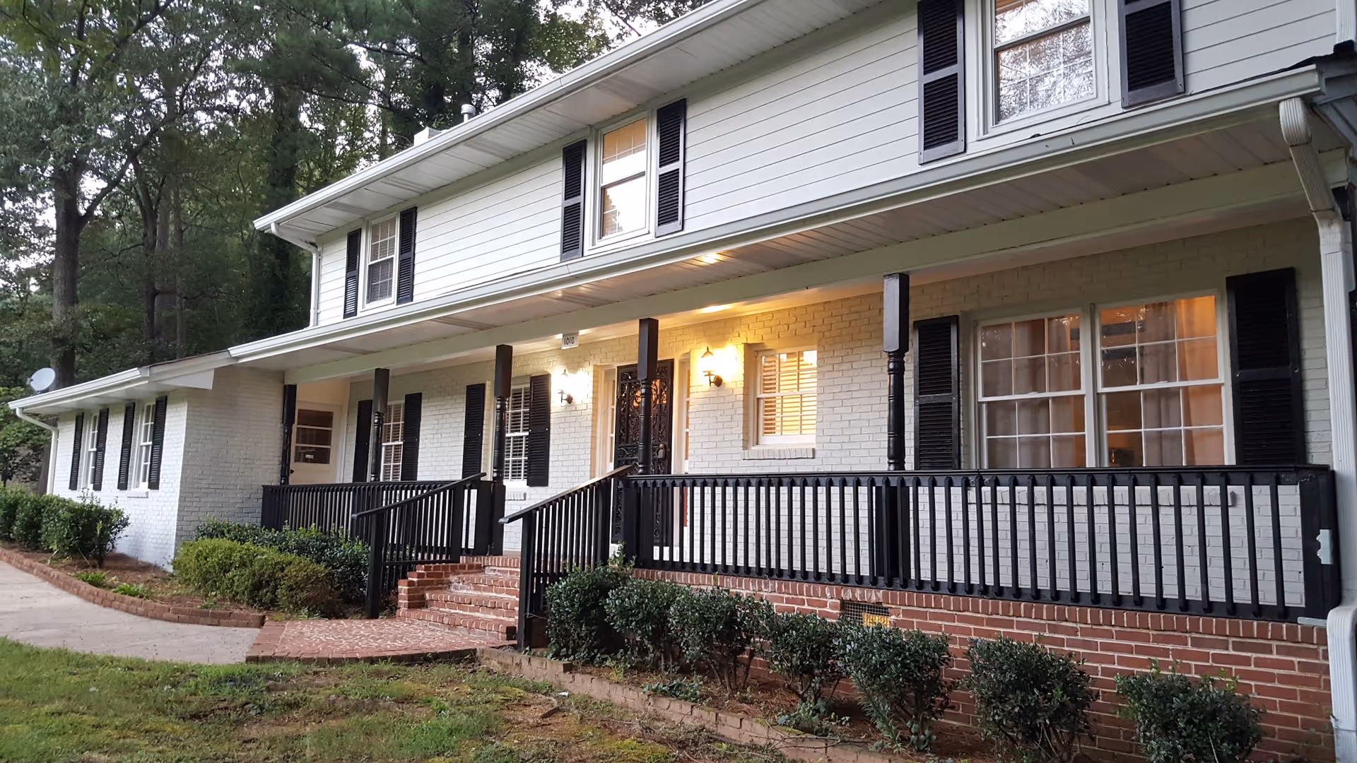Exterior view of a two-story white brick building with black shutters and a black railing along a front porch with steps. The building is surrounded by greenery and trees.