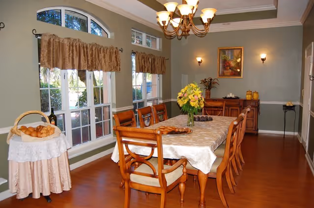 Formal dining room with a long wooden table and chairs, chandelier, floral centerpiece, sideboard, and large windows with valances.