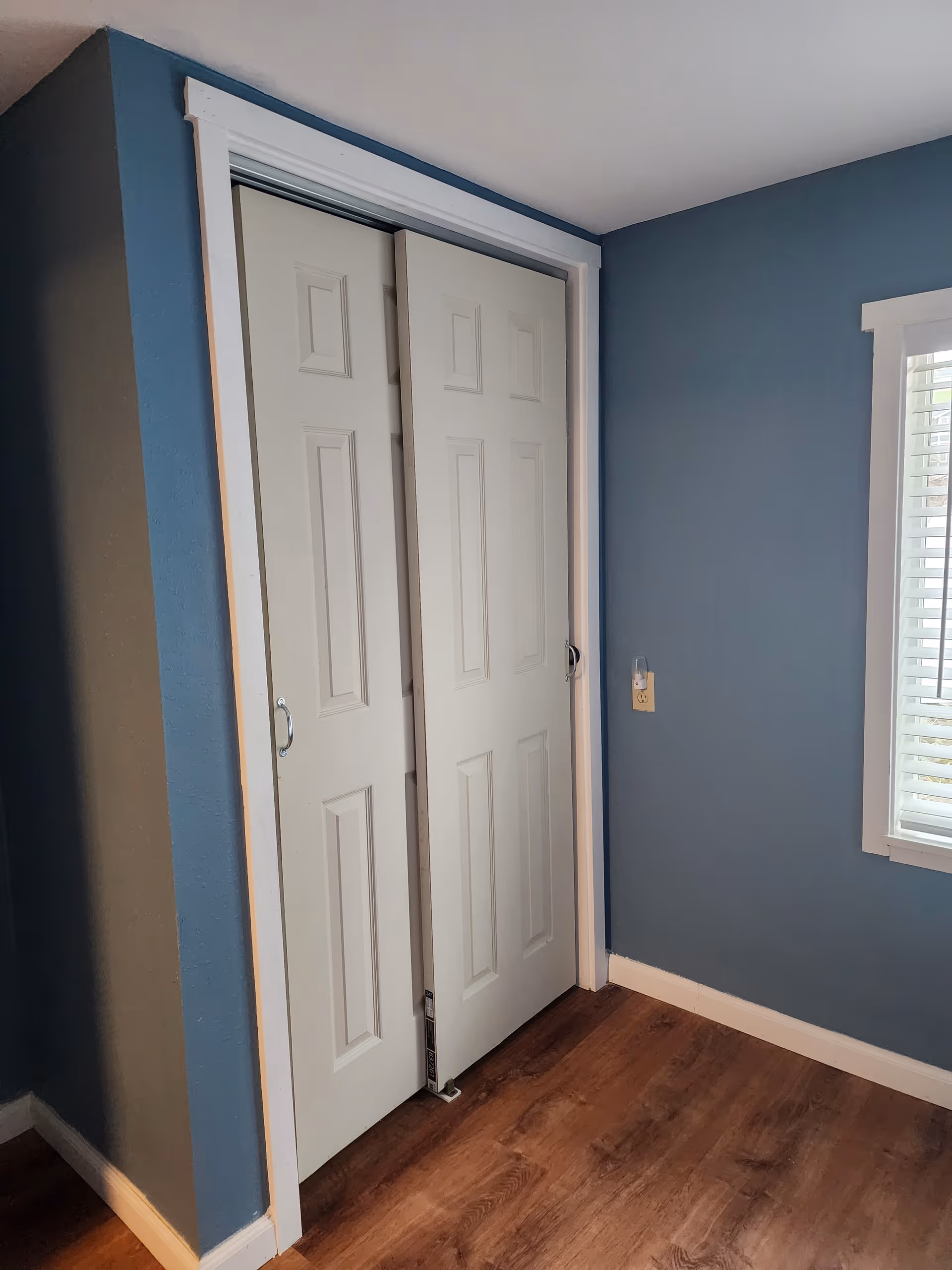 Interior corner of a room with blue walls, white trim, and wood flooring. There is a white sliding closet door and a window with white blinds on the right wall.