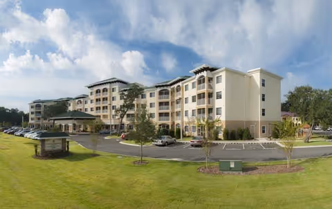 A multi-story senior living facility with a curved driveway, cars, and green lawn.