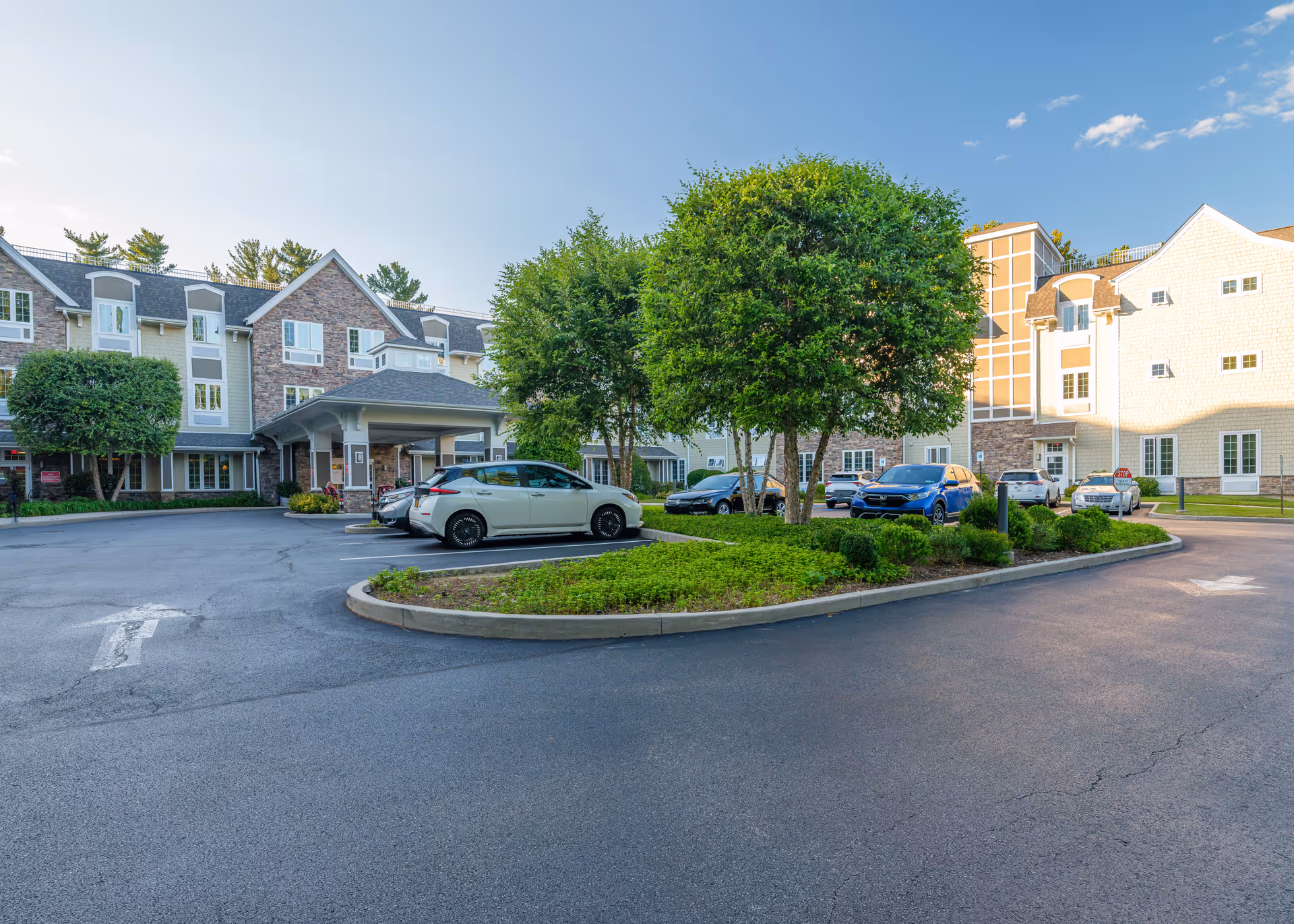 Exterior view of The Ambassador of Scarsdale senior living facility showing a parking lot with several cars, landscaped greenery with trees, and a multi-story building with a covered entrance under a clear blue sky.