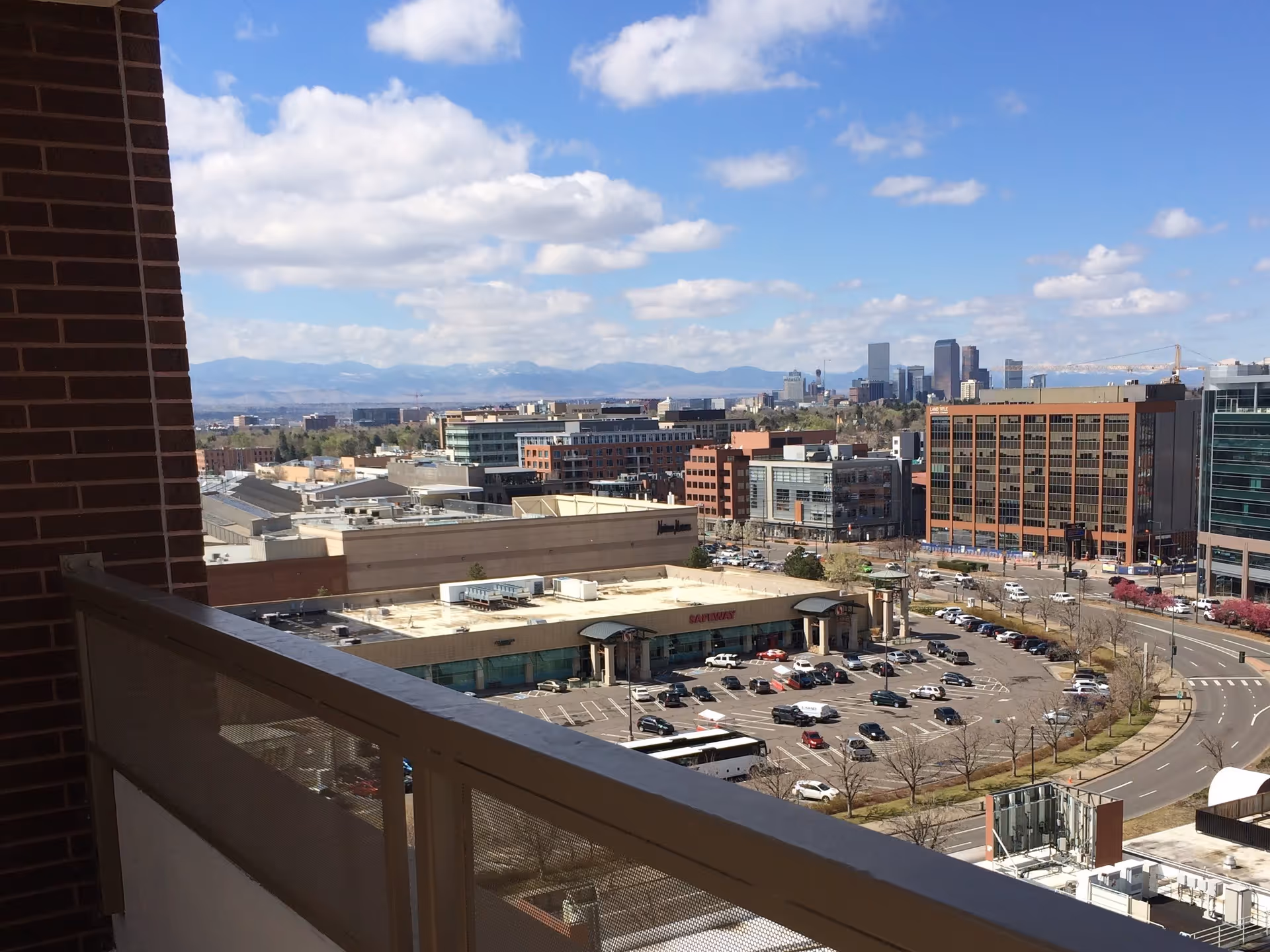 View from a balcony overlooking a parking lot with cars, a Safeway store, and multiple commercial buildings in the background under a partly cloudy sky with mountains in the distance.