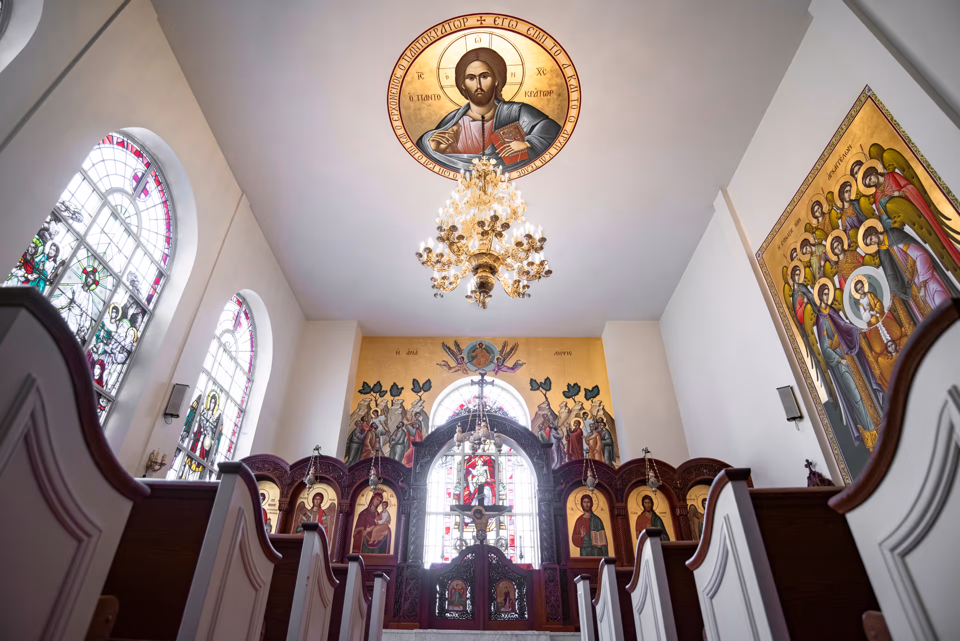 Interior view of a chapel with wooden pews, stained glass windows, religious icons, and a large chandelier hanging from the ceiling. The ceiling features a circular painting of Jesus Christ, and the walls are adorned with religious murals and iconography.