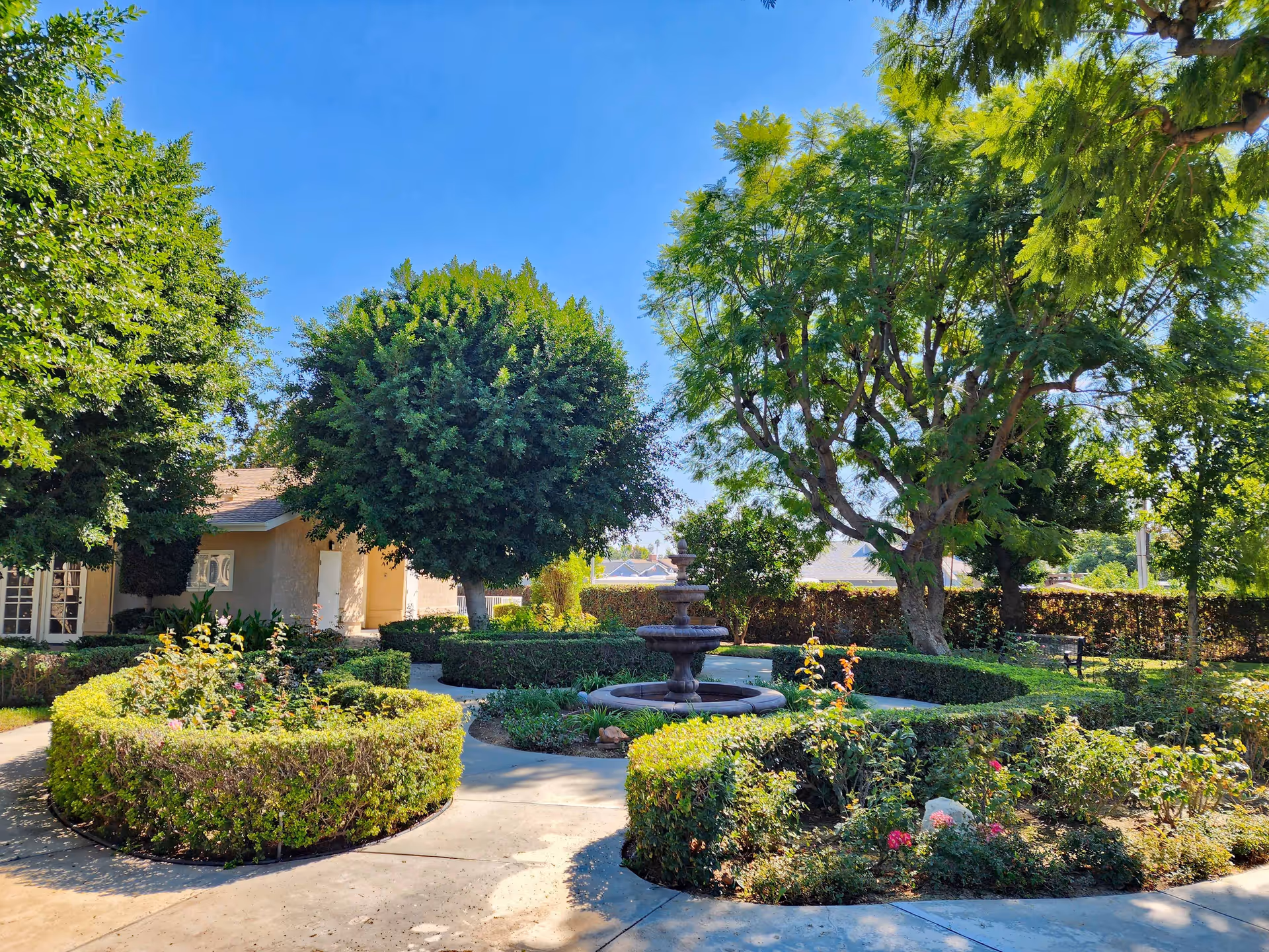 A sunny outdoor garden area with neatly trimmed hedges, a central tiered water fountain, various trees providing shade, and a beige building partially visible in the background under a clear blue sky.